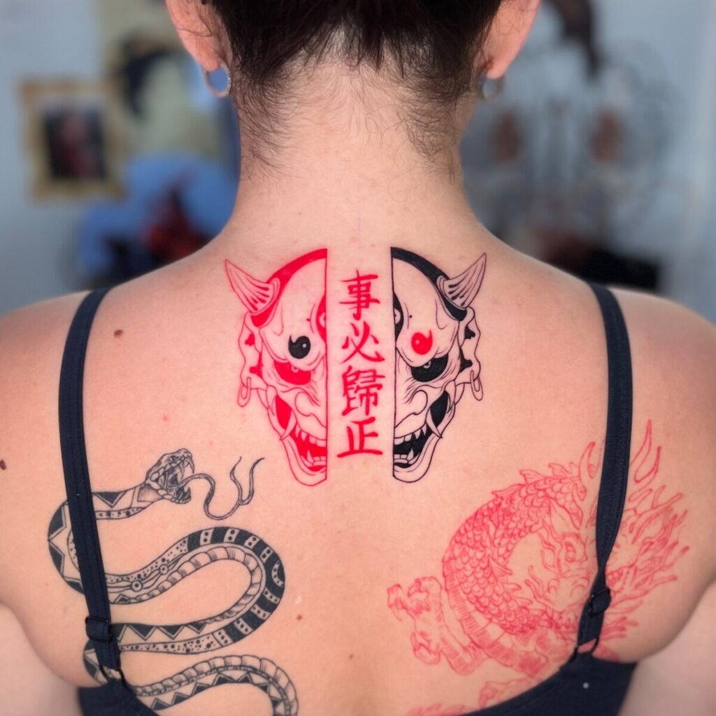 A bold tattoo covers a person's upper back, featuring traditional Japanese imagery. Two koi fish, one red and one outlined, appear to be swimming in a circular motion, resembling the yin and yang symbol. They are complemented by a red Hannya mask, signifying a jealous female demon in Noh theatre, and Japanese kanji characters in the center that add an element of depth. Intricate scales on the fish and detailed expression on the mask show a high level of craftsmanship. The person's hair is tied up, fully revealing the artwork, and the dark tank top straps contrast with the vibrant red ink.